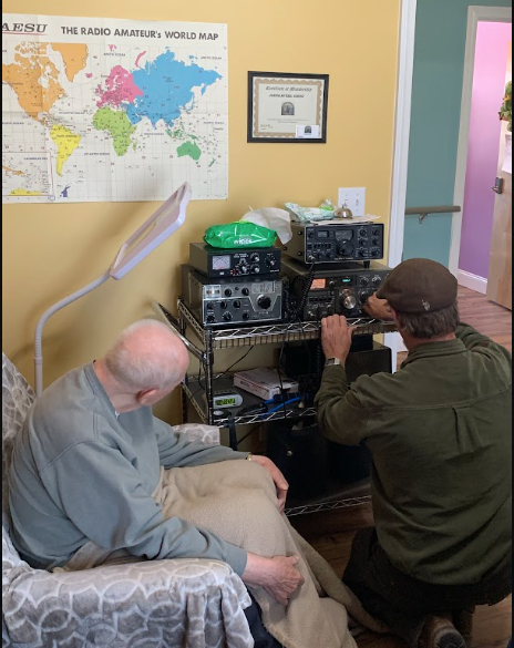 A retired Air Force resident in his room at Lavender Fields with his HAM radio equipment, world map, and amateur radio certificate on the wall